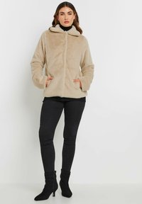 Beige falsk pelsfrakke med hætte, knaplukning og to sidelommer. Båret over en sort turtleneck, parret med mørke jeans og ankelstøvler.