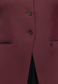 Blazer bordeaux à deux boutons, deux poches fendues horizontales et doublure intérieure noire visible à travers la fente avant.