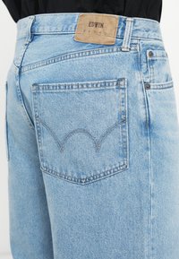 Jeans de denim azul claro com um remendo de pele castanha rotulado "EDWIN". Apresenta dois bolsos traseiros com detalhe de costura curva.