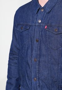 Giacca di jeans di colore blu scuro con chiusura a bottoni, due tasche sul petto e bottoni metallici. Tessuto testurizzato con dettagli di cucitura visibili.