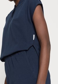 Dark blue short-sleeve jumpsuit with a high neckline and gathered elastic waistband. Features a small brand label on the side seam.