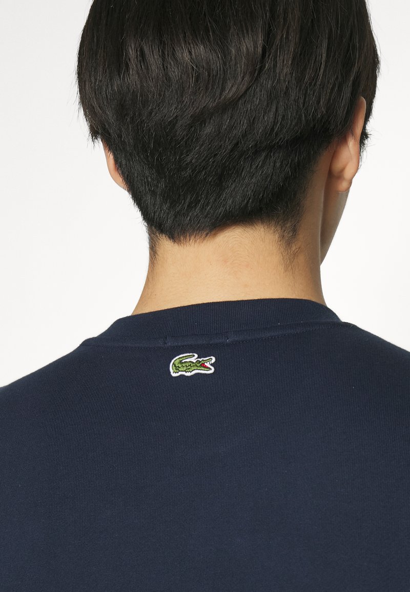 Granatowy bawełniany sweatshirt z małym zielonym logo aligatora wyszytym na tylnej części szyi. Gładka tekstura i casualowy styl.