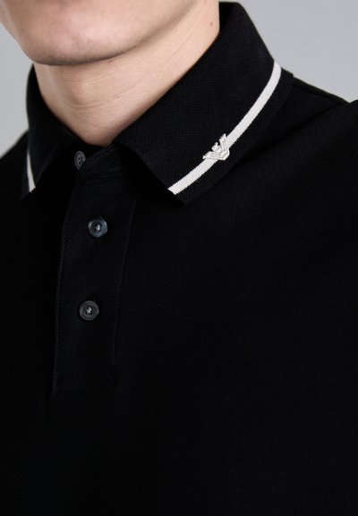 Black polo shirt with a textured fabric, three black buttons, and a collar featuring white stripes and a small embroidered logo.