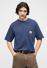 Νέος άνδρας με μεσαίου μήκους σκούρα μαλλιά, φοράει μπλε μπλούζα με τσέπες, καφέ παντελόνι και μαύρη ζώνη, στέκεται με τα χέρια στις τσέπες.