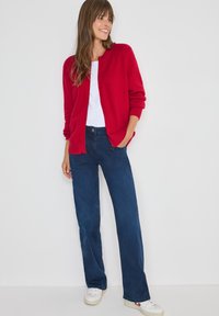 Cárdigan de punto rojo sobre una camiseta blanca, combinado con unos vaqueros de pierna ancha azul oscuro y zapatillas blancas. Presenta puños y dobladillo acanalados.