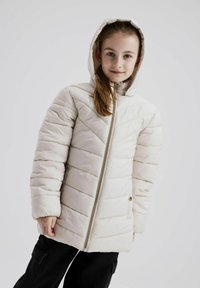 Chaqueta acolchada de color beige claro con capucha, presenta un patrón de acolchado diagonal, cremallera dorada y bolsillos laterales. Llevada con pantalones negros.