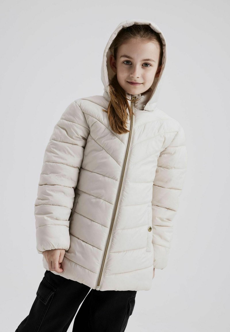 Chaqueta acolchada de color beige claro con capucha, presenta un patrón de acolchado diagonal, cremallera dorada y bolsillos laterales. Llevada con pantalones negros.