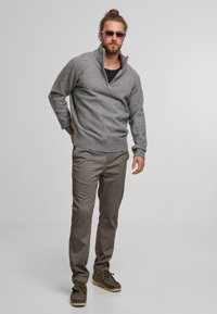 Gray knit sweater with a half-zip collar, paired with brown plaid trousers and olive green suede shoes. Clear sunglasses complete the look.