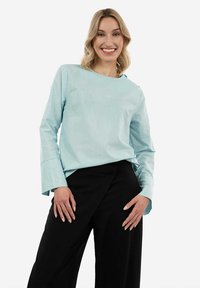 Top azzurro chiaro a maniche lunghe con finitura testurizzata, scollatura rotonda e ampi polsini, abbinato a pantaloni neri a gamba larga. Design semplice.