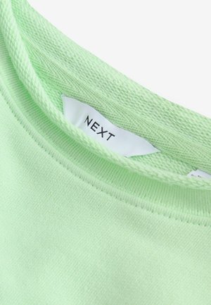 T-shirt en coton vert clair avec un col rond et un détail en tricot texturé, arborant une étiquette blanche avec le nom de la marque "NEXT" à l'intérieur.