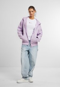 Haut de sweat à capuche zippé violet clair avec poches avant, porté sur un t-shirt blanc avec un petit motif noir, associé à un jean bleu clair ample et des baskets blanches.