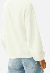 Bílá mikina s volným střihem, spadlými rameny a žebrovanými manžetami, ve spojení se světle modrými zmáčknutými denimovými šortkami.