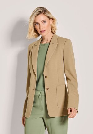 Beige textuurblazer met twee gouden knopen, een enkele borstzak en gelapte revers, gecombineerd met een groen topje en bijpassende groene broek.