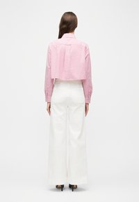 Γυναίκα που στέκεται με την πλάτη στραμμένη, φορώντας ένα κοντό ροζ και λευκό ριγέ πουκάμισο, λευκό παντελόνι με φαρδιά μπατζάκια κα ι μαύρα ψηλοτάκουνα.