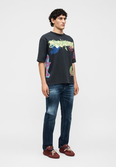 Άνδρας όρθιος, φοράει μαύρο γραφικό T-shirt, ξεβαμμένο μπλε τζιν και κόκκινα αθλητικά με μπεζ κορδόνια, σε απλό γκρι φόντο.