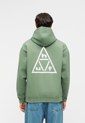 Mand iført en grøn hættetrøje med et hvidt trekantet logo, der indeholder bogstaverne h, u og f på ryggen, kombineret med blå jeans.