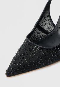 Pantof negru cu vârf ascuțit, confecționat dintr-un material texturat, împodobit cu nituri negre strălucitoare, având o panou lateral din plasă transparentă și o căptușeală interioară netedă.