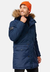Parka bleu marine isolée avec capuche en fourrure, devant boutonné et deux poches avant. Comprend des accessoires noirs et une finition en tissu texturé.