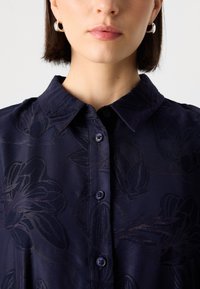 Donkerblauwe shirt met een bloemenjacquardpatroon, voorzien van een klassieke kraag, knoopsluiting en ronde blauwe knopen. Soepele textuur.