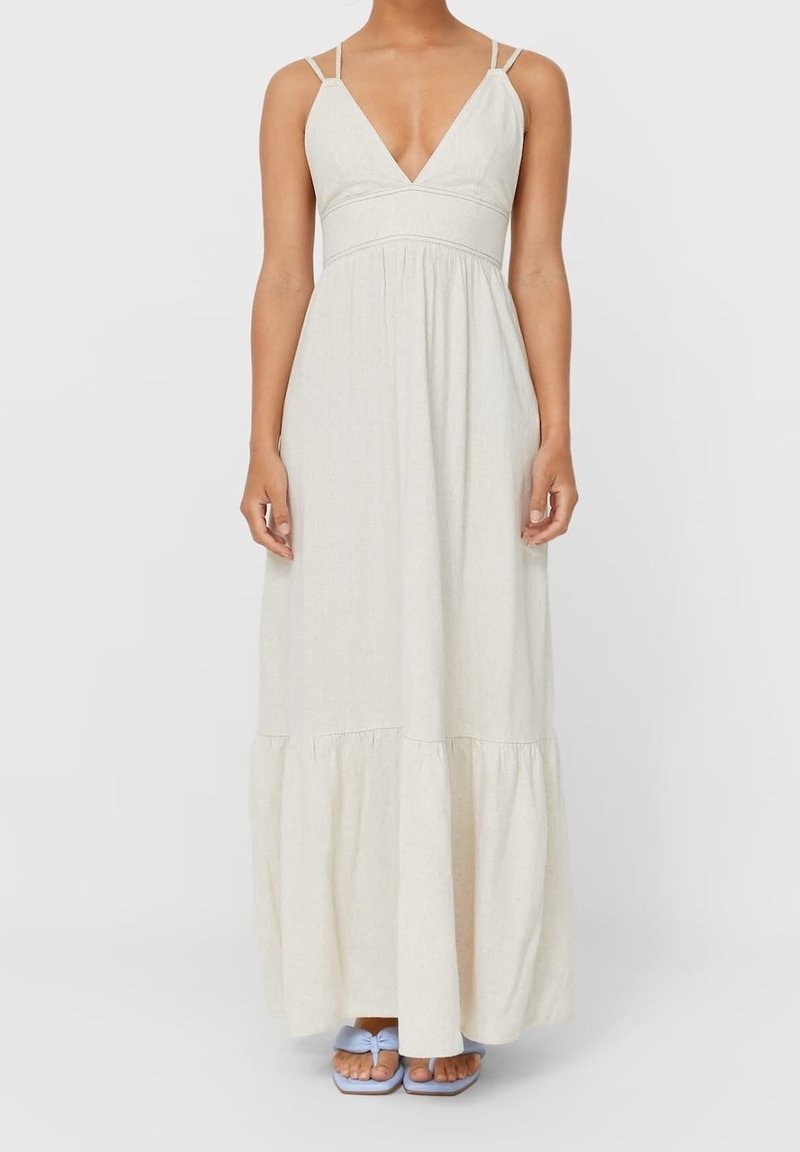 Femme portant une robe sans manches beige clair, longue jusqu'au sol, avec un décolleté plongeant en V et des bretelles doubles, assortie à des sandales ouvertes bleu clair.