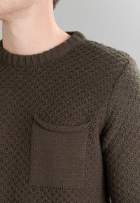 Maglione lavorato a maglia verde oliva con un motivo testurizzato, scollo rotondo e una tasca applicata frontale. Presenta polsini e orlo a costine.