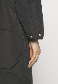 Abrigo negro acolchado con un diseño acolchado, que muestra un primer plano de la manga y el detalle del botón a presión, textura suave y forma ajustada.