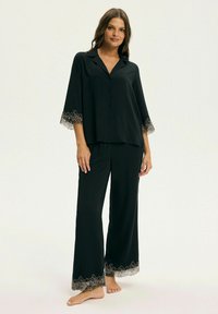 Kobieta stoi boso, ubrana w czysty, luźny zestaw piżamy w kolorze czarnym z koronkowymi wykończeniami na rękawach i nogawkach, na tle jasnym i jednolitym.