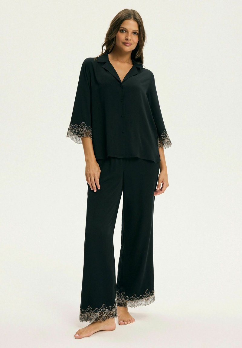 Kobieta stoi boso, ubrana w czysty, luźny zestaw piżamy w kolorze czarnym z koronkowymi wykończeniami na rękawach i nogawkach, na tle jasnym i jednolitym.