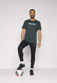 Tumši zaļš krekls ar uzrakstu "LONDON" un lauvas grafiku, apvienots ar melnajām sporta biksēm un melnajām futbola kurpēm, kāja atpūšas uz futbola bumbas.