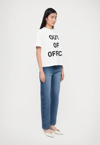 Λευκό cropped t-shirt με το μήνυμα "OUT OF OFFICE" τυπωμένο με έντονα μαύρα γράμματα, συνδυασμένο με μπλε τζιν παντελόνι με ευθεία γραμμή και λευκά μπαλαρίνια με μυτερή μύτη.