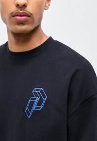 Gros plan sur une personne portant un sweat-shirt noir avec un design de cube géométrique bleu sur le côté gauche de la poitrine, sur un fond clair.