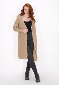 Pletený béžový cardigan s texturovaným vzorem, dlouhými rukávy a několika knoflíky vpředu. V kombinaci s černými těsnými kalhotami a kotníkovými botami.