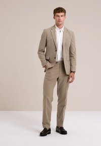 Homme debout vêtu d'un costume beige avec une chemise blanche et des chaussures noires devant un fond beige uni.