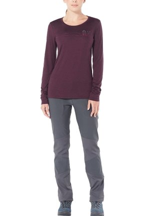 Mujer con camiseta de manga larga color granate, pantalones grises para exteriores con rodillas reforzadas y botas de senderismo azules, de pie con los brazos relajados.