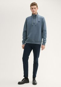 Sweatshirt bleu marine à col, en tissu texturé, avec un système de réglage par cordon de serrage et un petit logo. Associé à un pantalon sombre et des chaussures noires à lacets.