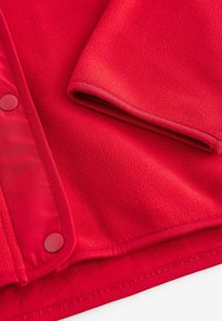 Rode fleece jasje met een soepele textuur, knoopsluitingen en een zachte binnenvoering. Accenten zijn onder andere gestikte zomen en een naadloos ontwerp.