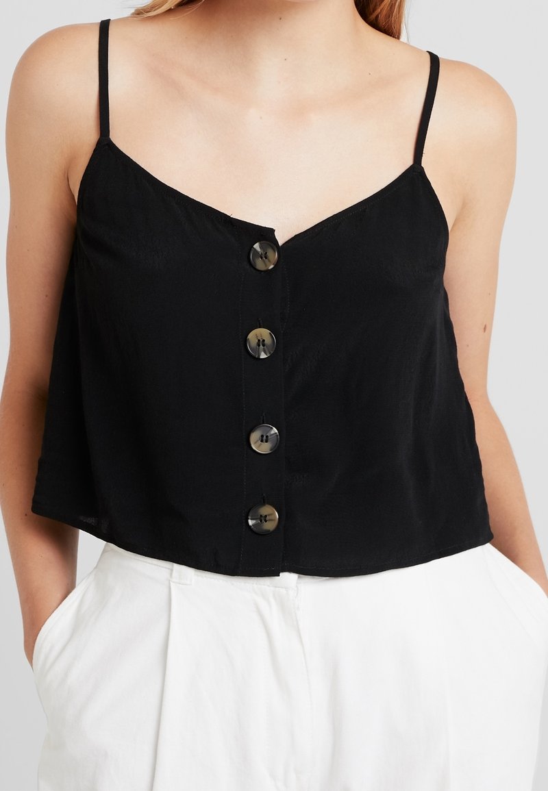 Top court sans manches noir avec un décolleté en forme de V et cinq boutons à l'avant. Le tissu a une texture douce, associé à un pantalon blanc taille haute.