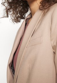 Blazer marrone chiaro con una texture liscia e una tasca unica, indossato sopra un top bordeaux. Vista ravvicinata, con attenzione ai dettagli del tessuto e del design.