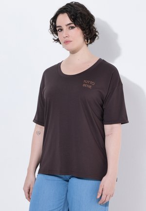 Frau mit kurzen lockigen Haaren, die ein braunes T-Shirt mit dem Schriftzug „TUTTO BENE“ und hellblaue Jeans trägt, steht vor einer weißen Wand.