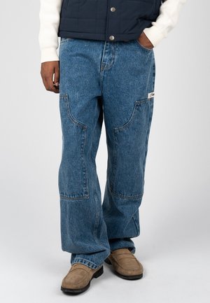 Mann trägt lose sitzende blaue Denim-Jeans mit großen Pattentaschen und hellbraunen Wildleder-Loafers auf hellem Hintergrund.