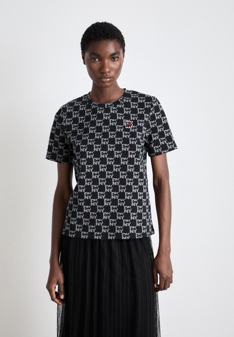 Μαύρο κοντομάνικο μπλουζάκι με επαναλαμβανόμενο γραφικό μοτίβο "NY", που διαθέτει μια μικρή κόκκινη ετικέτα λογότυπου στο στήθος. Φοριέται με μια μαύρη πτυχωτή φούστα.