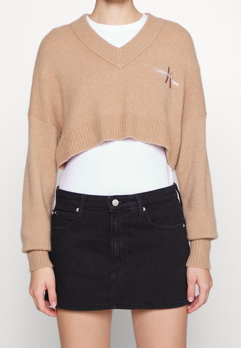 Pull beige court à col en V porté sur une chemise blanche, associé à une mini jupe en denim noir avec poches avant et fermeture boutonnée.