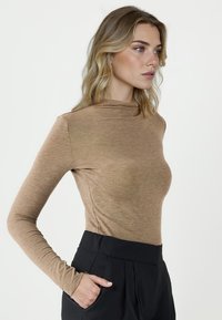 Długi rękaw, dopasowana bluzka w jasnobrązowym, miękkim materiale z wysokim dekoltem i bezszwowymi rękawami, w połączeniu z czarnymi spodniami o wysokim stanie.