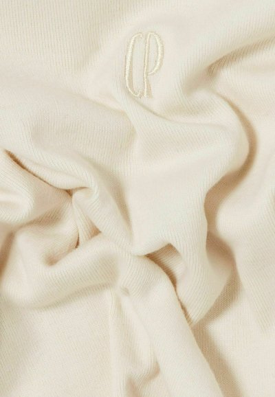 Tessuto a maglia color crema con sottili pieghe e un logo ricamato "CP" in filo coordinato vicino al centro.
