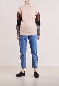 Rosa hoodie med mönstrade svarta, bruna och grå ärmar, tillsammans med blå denimjeans och svarta slip-on skor.