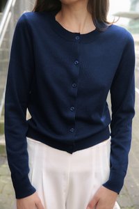 Marineblaue Strickjacke aus geripptem Strickstoff, mit rundem Halsausschnitt, Knopfverschluss und langen Ärmeln. Über hellen Hosen getragen.