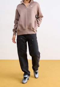 Ljusbrun zip-up hoodie med ribbade manschetter, tillsammans med svarta baggybyxor med guld accenter och grå sneakers.