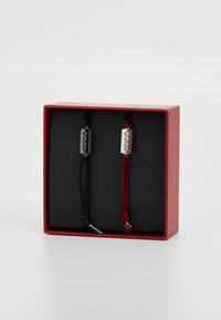 Deux bracelets en cordon tressé, l'un noir et l'autre rouge, avec des plaques métalliques gravées "HUGO" à l'intérieur d'une boîte-cadeau rouge.