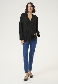 Schwarze Bluse mit langen Ärmeln, vertikalen silbernen Streifen, V-Ausschnitt und strukturiertem Stoff; kombiniert mit blauen Slim-Fit-Jeans und schwarzen spitzen Ballerinas.