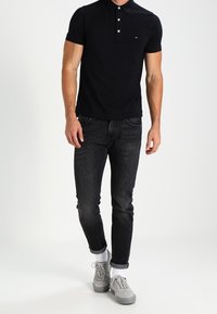 Svart polotröja med en krage med tre knappar och liten logotyp, i kombination med åtsittande mörka jeans och grå sneakers med uppvikta ärmar.